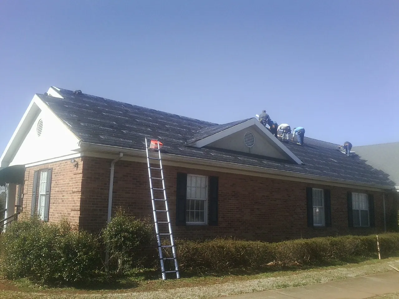 Crew tearing shingles off a brick ranch roof with a ladder set — Metal Roof Repair work by our crew in Drakesville