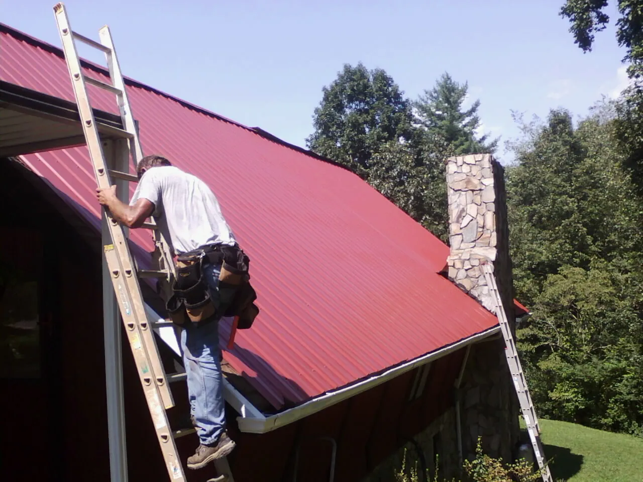 Roofer on a ladder beside a red metal roof and stone chimney — Soffit & Fascia Repair by Roofing Brothers in Drakesville